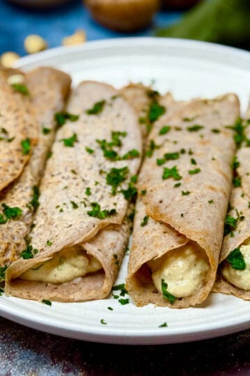 Four buckwheat crepes filled with a creamy mixture, garnished with chopped parsley, are arranged on a white plate. The background is softly blurred with hints of ingredients.