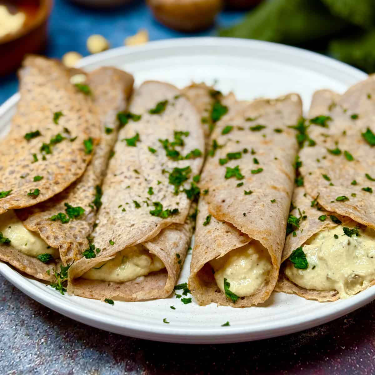 Four buckwheat crepes filled with a creamy mixture, garnished with chopped parsley, are arranged on a white plate. The background is softly blurred with hints of ingredients.