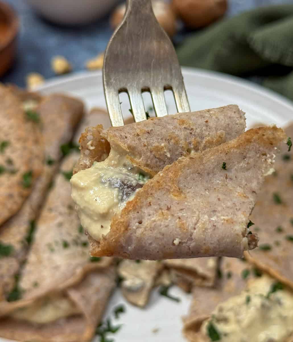 A close-up of a fork holding a buckwheat crepe filled with creamy mushroom filling, with more crepes garnished with chopped herbs visible on a plate in the background.