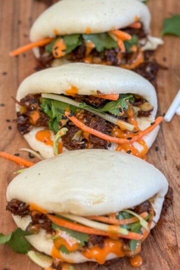 Three steamed bao buns filled with shredded meat, julienned carrots, fresh cilantro, and drizzled with an orange sauce are arranged on a wooden board, with a pair of chopsticks placed beside them.