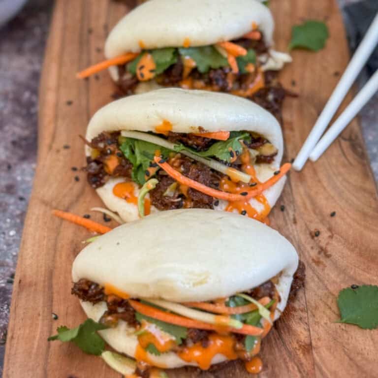 Three steamed bao buns filled with shredded meat, julienned carrots, fresh cilantro, and drizzled with an orange sauce are arranged on a wooden board, with a pair of chopsticks placed beside them.