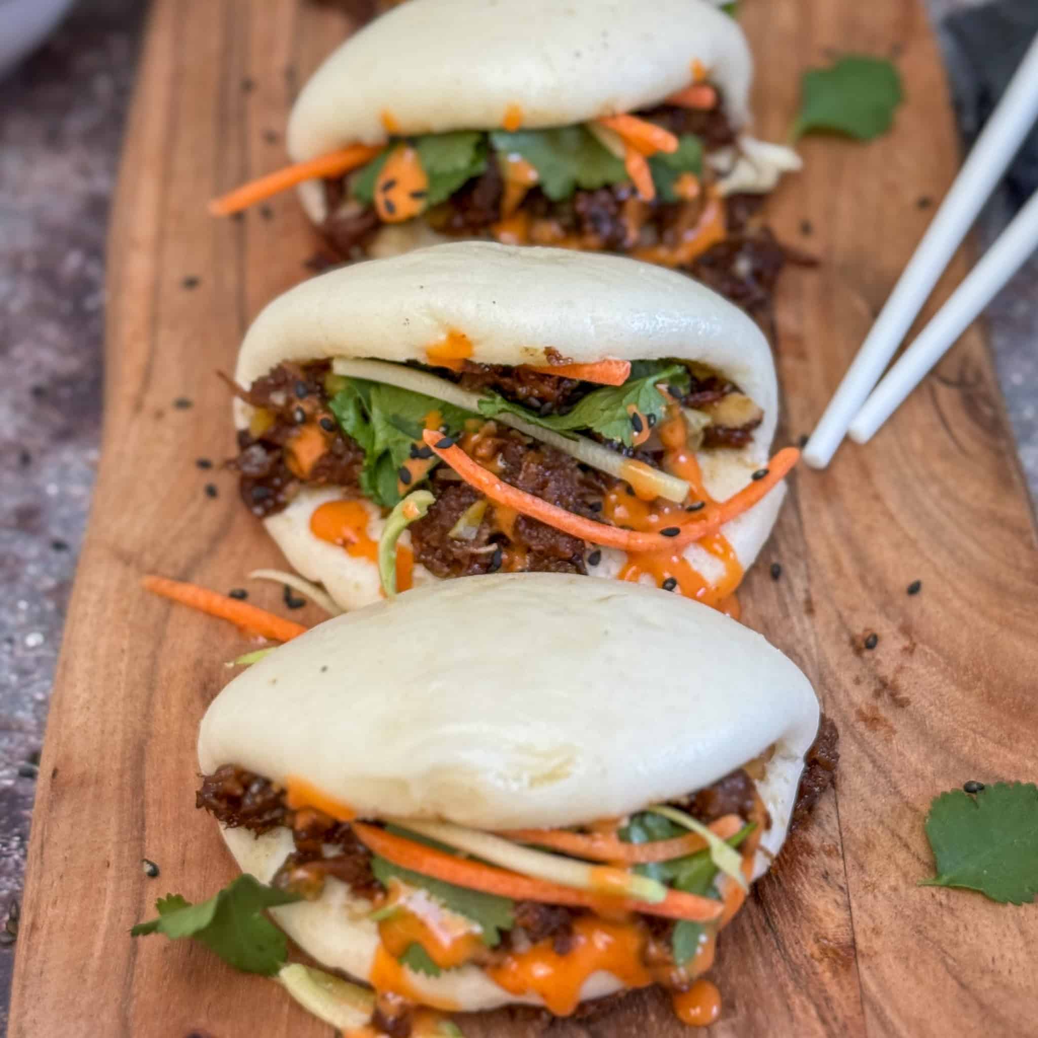 Three steamed bao buns filled with shredded meat, julienned carrots, fresh cilantro, and drizzled with orange sauce, are arranged on a wooden board with a pair of chopsticks beside them.