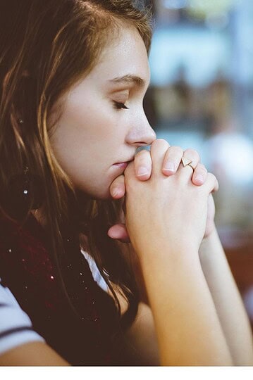 A young vegan woman sits with her eyes closed and hands clasped in front of her face, appearing deep in thought or prayer in a softly blurred indoor setting.