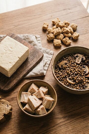 A wooden table with various plant-based protein sources, including a block of tofu on a cutting board, tempeh, seitan slices, soy protein chunks, lentils, and mushrooms in bowls.
