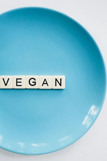 A blue plate with white letter tiles spelling VEGAN in the center, placed on a white surface. A green plant leaf is visible on the upper left edge of the plate.