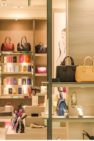 A brightly lit store display features handbags in black, beige, and blue on shelves, with various shoes, scarves, and boxed items arranged neatly below and behind them. The background shows more handbags and a blurred poster.