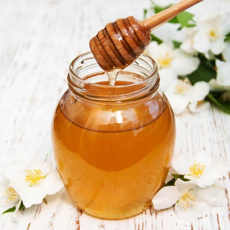 Fresh honey in a glass jar with a honey dipper on a white wooden surface.