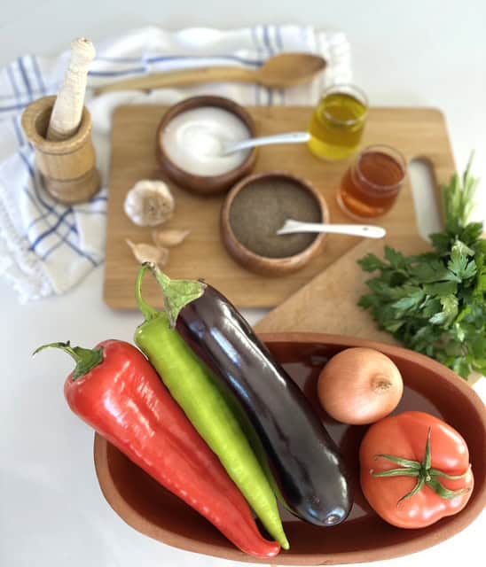 Fresh eggplant, red and green peppers, onion, and herbs on a wooden tray.